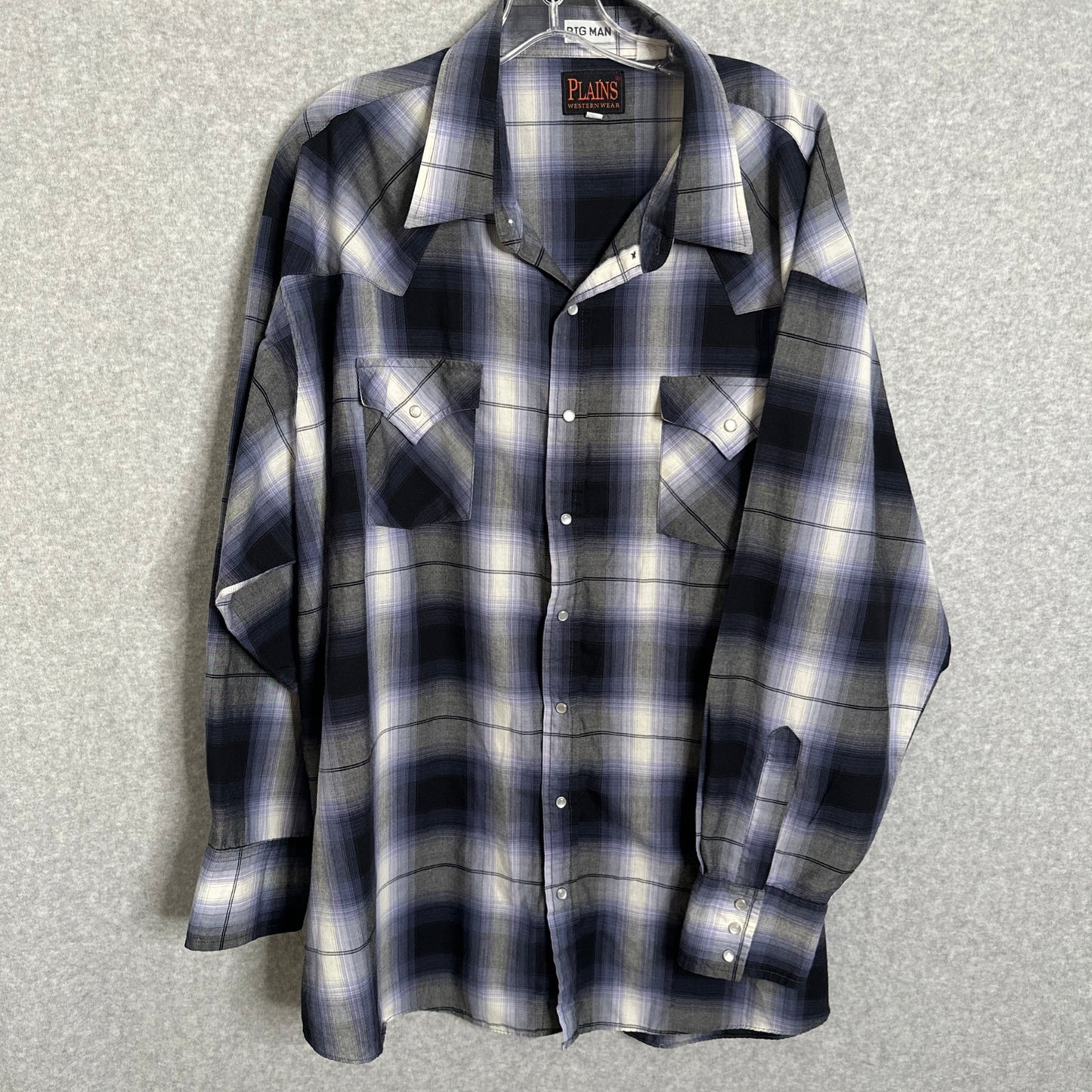 A long-sleeve plaid button-up shirt by Plains, featuring a navy, gray, and white checkered pattern, hanging on a hanger against a textured gray background.