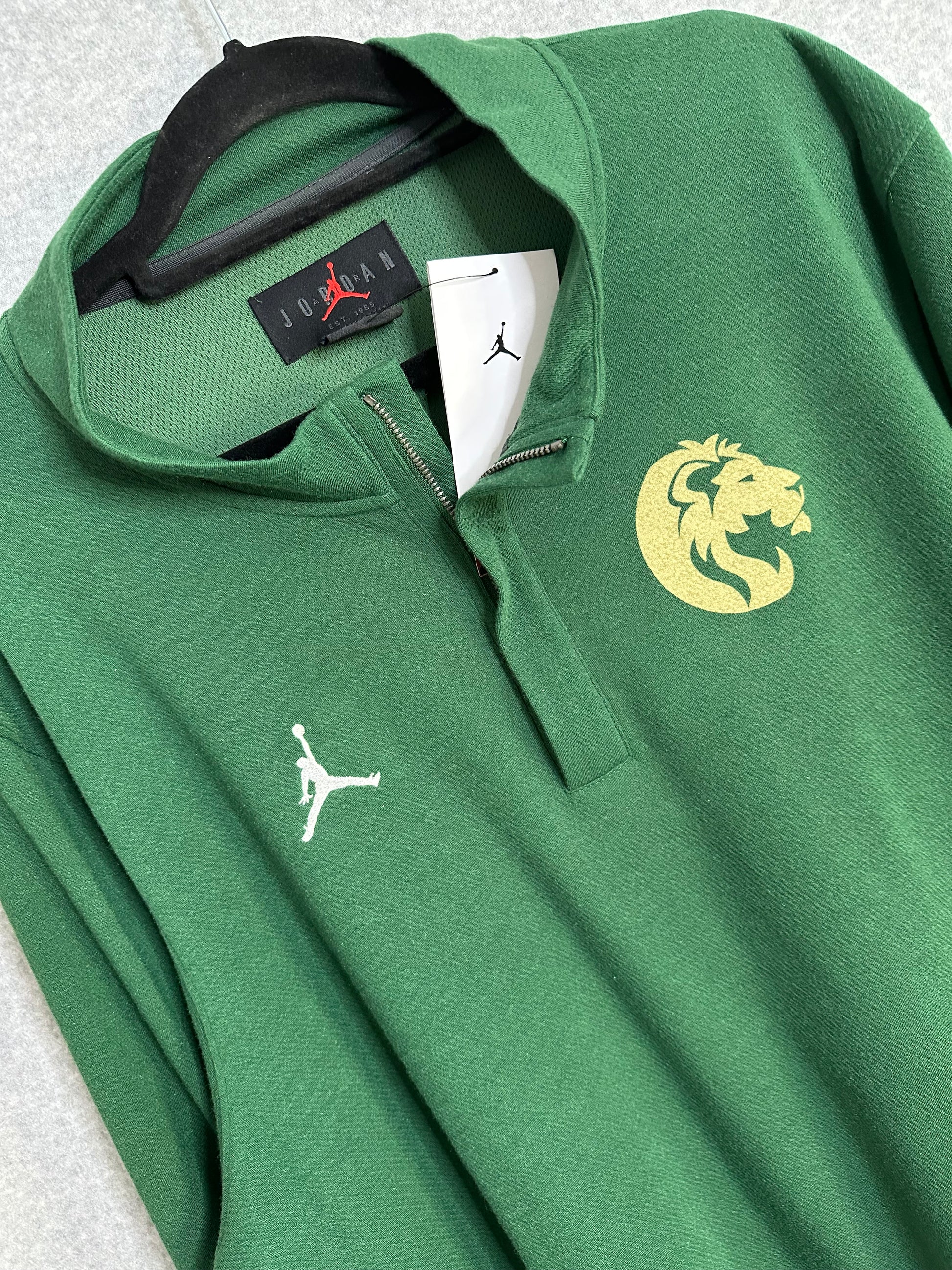 A green pullover sweatshirt with a half-zip collar, featuring a white embroidered Jumpman logo on the left chest and a gold embroidered lion head logo on the right chest. The sweatshirt is shown hanging on a black hanger with a white tag.