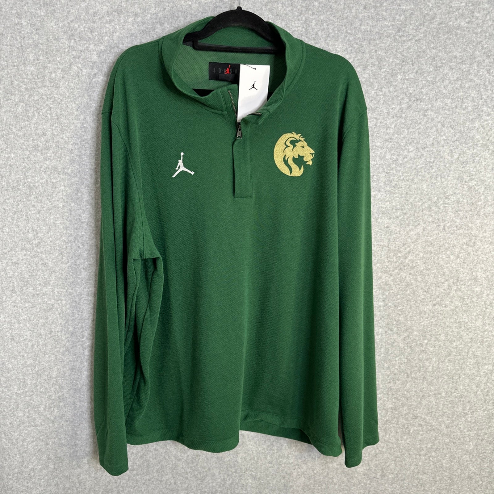 A green long-sleeve quarter-zip pullover sweatshirt by Jordan, featuring a white Jumpman logo on the right chest and a gold lion head logo on the left chest, hanging on a black hanger against a gray textured background.