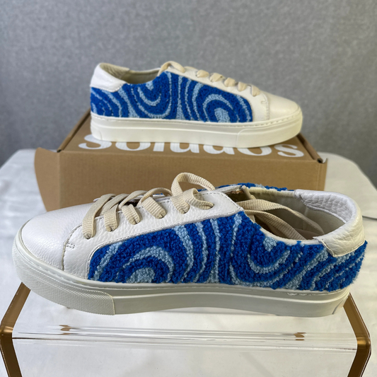 White sneakers with blue pattern on a glass surface, with a Soludos box in the background.