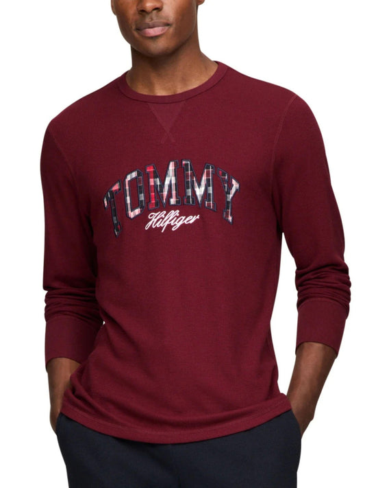 A man wearing a long-sleeve crewneck sweatshirt in maroon with a graphic 'TOMMY Hilfiger' logo on the chest, set against a plain white background.